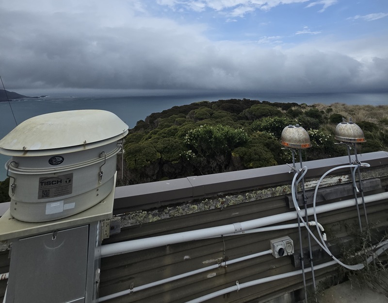 A metallic dome-shaped machine with two nearby antennae sits nearby lush green forestry near the coastline.