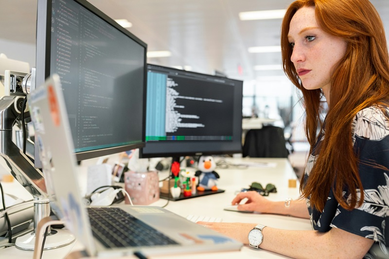 A woman with long red hair is focused on coding at her desk, surrounded by multiple monitors displaying lines of code.