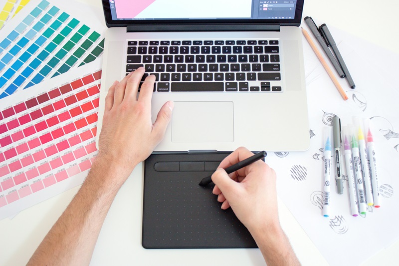 A designer's workspace featuring a laptop, a digital drawing tablet, and color swatches.