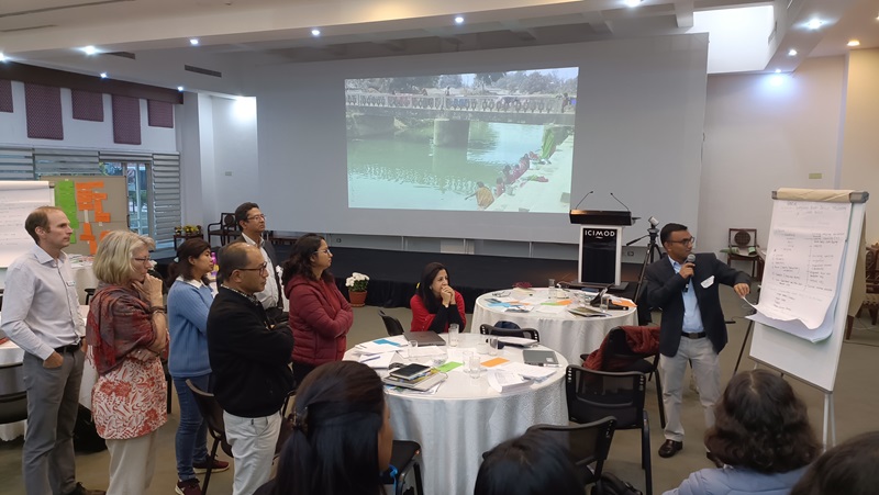 A group of people gather around a table whilst a man presents at a flipchart