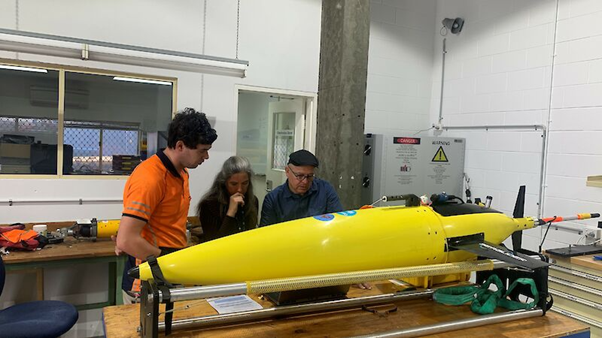 Three people inspect a yellow sea glider in a workshop.