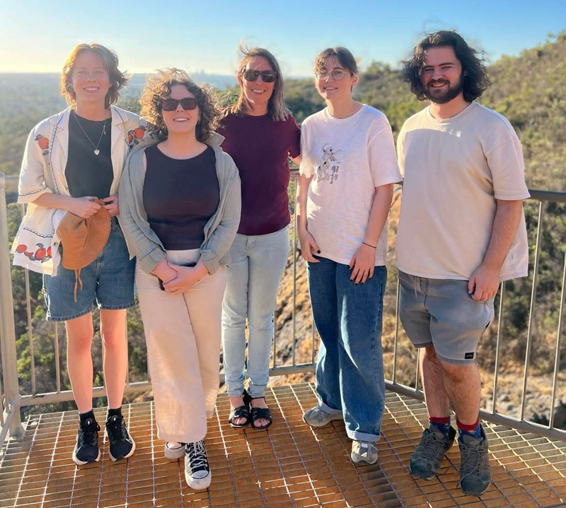 A group of five people pose for a photo on a grated deck with a hilly landscape and a setting sun in the background.