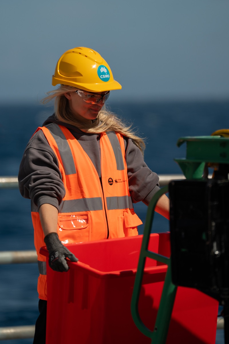 A person in a hi vis vest and yellow hard hat caryying a red tub on a ship at sea.