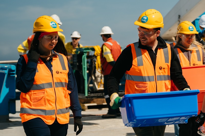 Two people in hi vis and hard hats walking across the deck of a ship at sea.