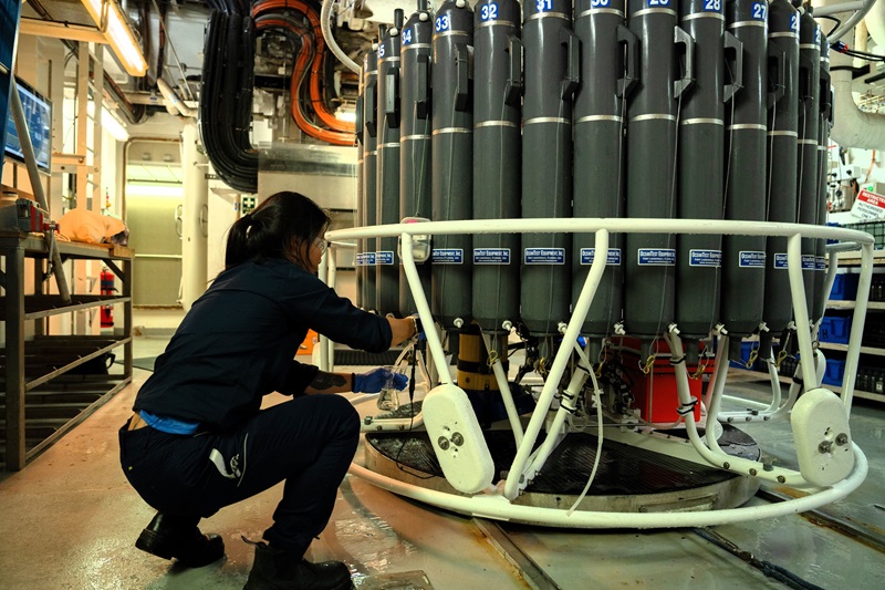 A person kneels next to a large circular piece of scientific equipment covered in long grey water bottles.