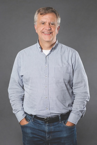 A man in a grey business shirt and jeans smiles at the camera with hands in his pockets.