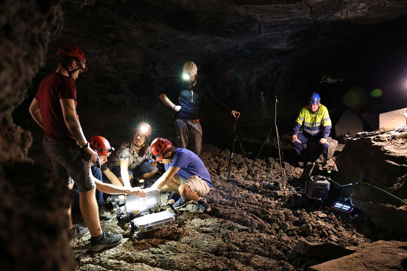 a group of people surround a robot on the ground spot lit by their head lamps inside a cave. 