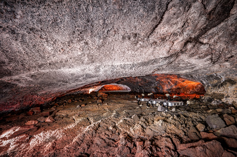 a segmented robot with curved paddle wheels, travelling away from the camera into a tunnel through rock with rough rocky floor and low-hanging ceiling. 