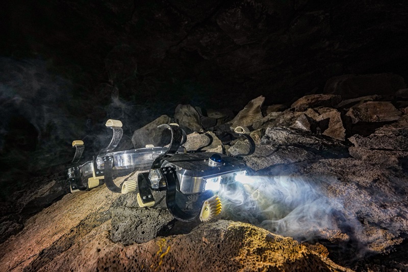 segmented rover with six curved paddles that work as wheels is moving over an uneven rocky surface inside a cave. two 'headlights' illuminate the rock in front of the rover, picking up a whisp of smoke in the air.