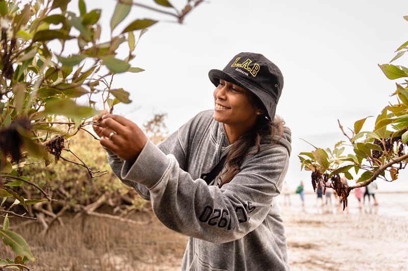 A young woman smiles while inspecting leaves on a tree branch near a beach, with blurred figures and a sandy shoreline in the background.