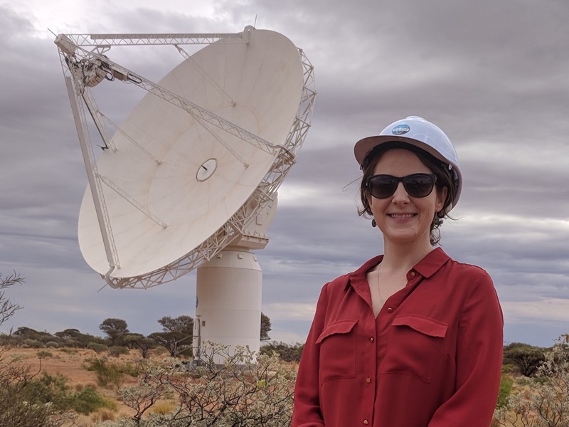 Elizabeth wearing a hard hat and smiling at the camera. Behind her is a large telescope dish antenna.