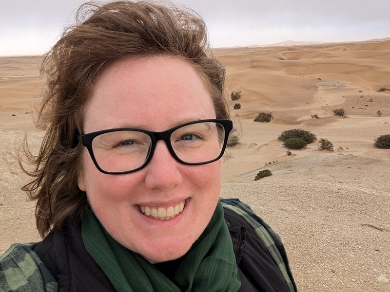 Smiling Becki stands in front of a vast desert landscape in Namibia, with sand dunes and sparse vegetation in the background.