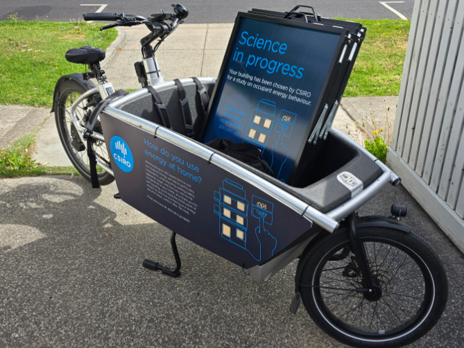 A cargo bike with a CSIRO logo on the side is parked on a footpath. The bike's cargo area contains a display board with the text "Science in progress," and information about a study on energy use at home.